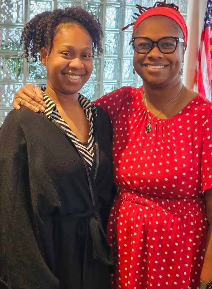 Two women smiling together indoors, one in black and one in red dress.